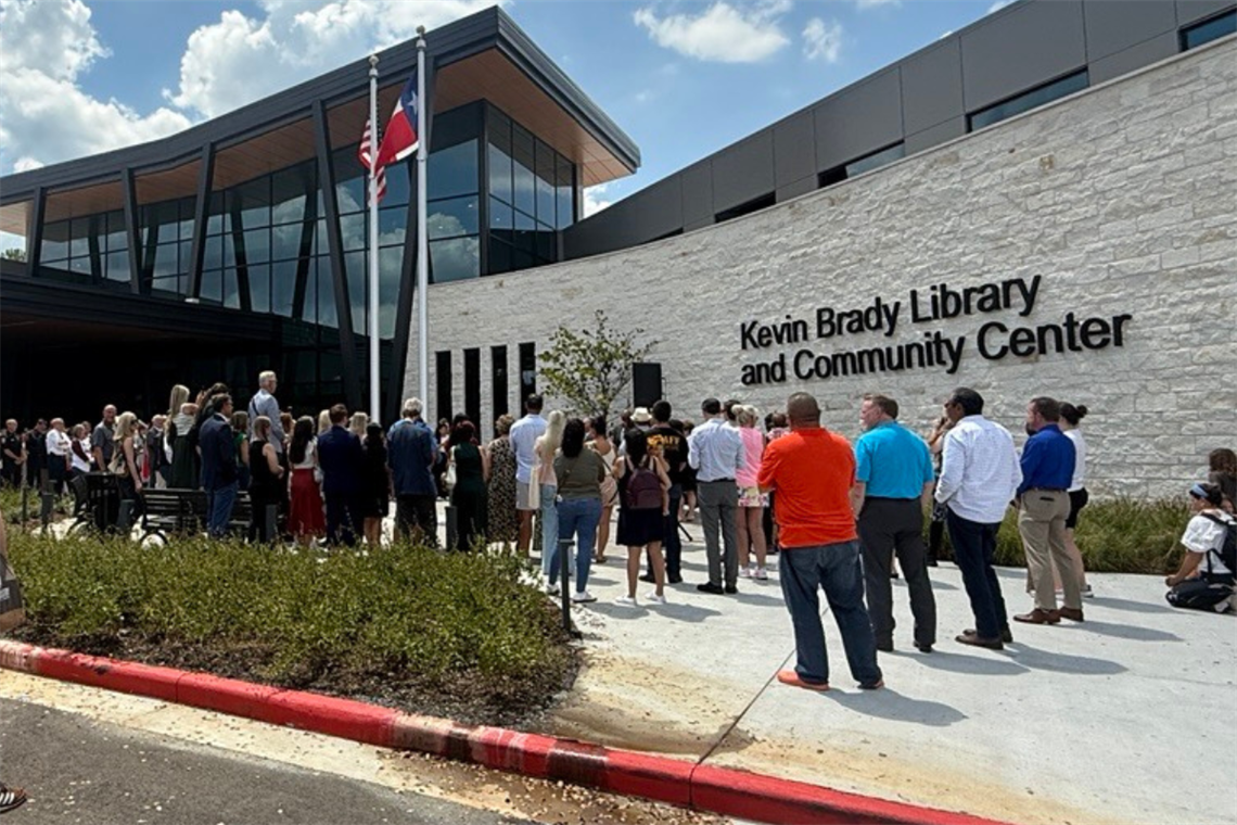 Photo of a white stone building, Kevin Brady Library and Community Center, with people standing around the entrance with 