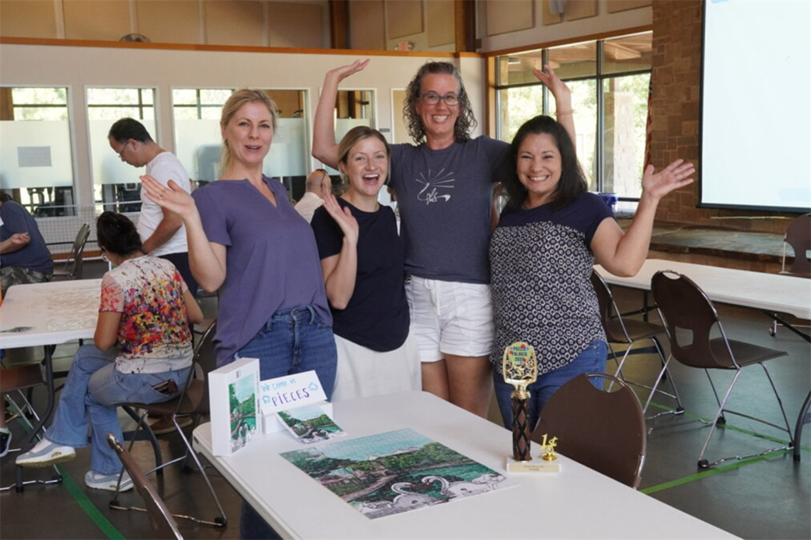 Photo of five smiling people waving in joy standing in front of a completed puzzle