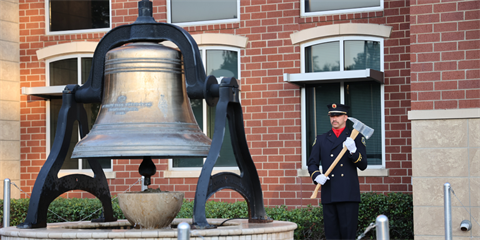 A uniformed firefighter stands beside a large historic bell, holding a ceremonial axe, in front of a brick building.