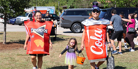 A family dressed in Halloween costumes as an oversized Doritos' bag and a can of Cola costumes walks holding the hand with a young girl in a purple skirt.