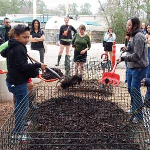 Class participants using various tools to turn an outdoor compost bin while others observe 