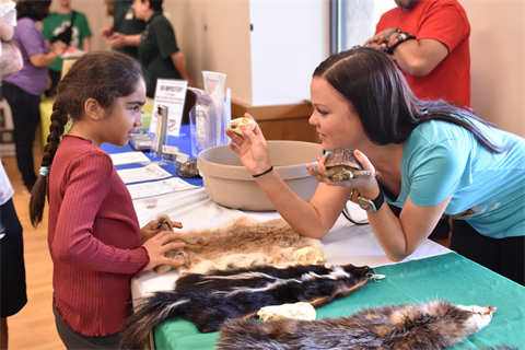 A woman leaning over a table with furs while holding a turtle and talking to a young girl