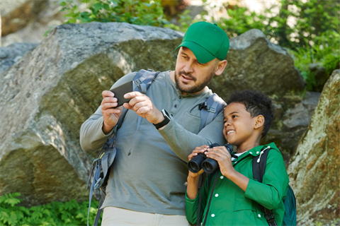 A child with a father outdoors appearing to identify species using a phone and binoculars