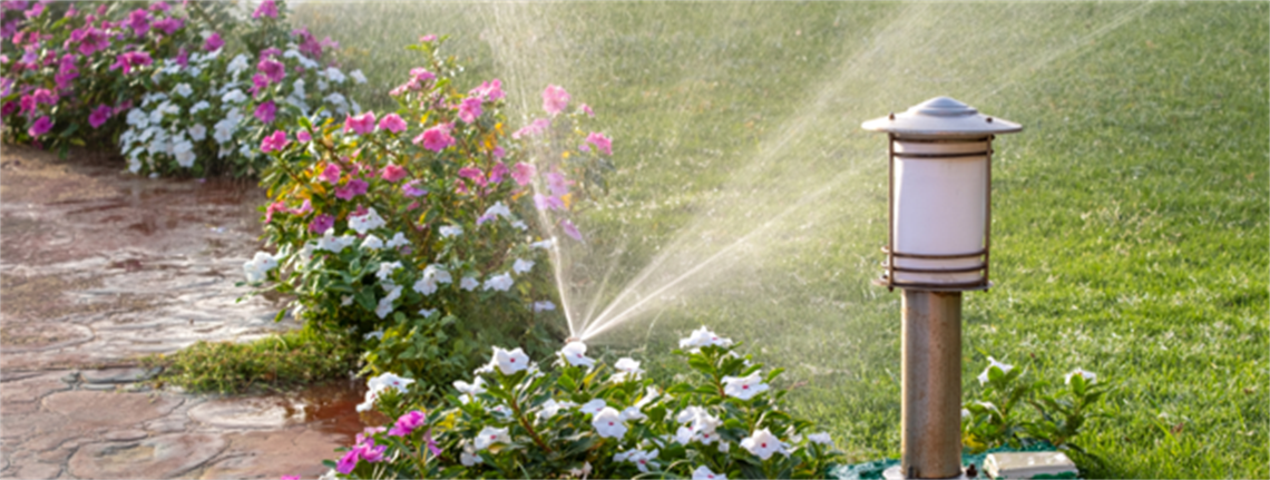 A sprinkler spraying water on grass and flowers next to a lamp