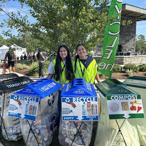 Two girls standing behind trash, recycling, and compost containers outside