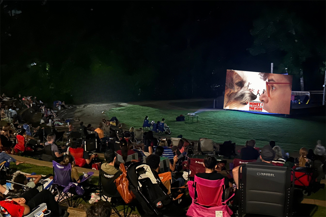 Families sitting on grass at a park watching an outdoor movie on a large screen.
