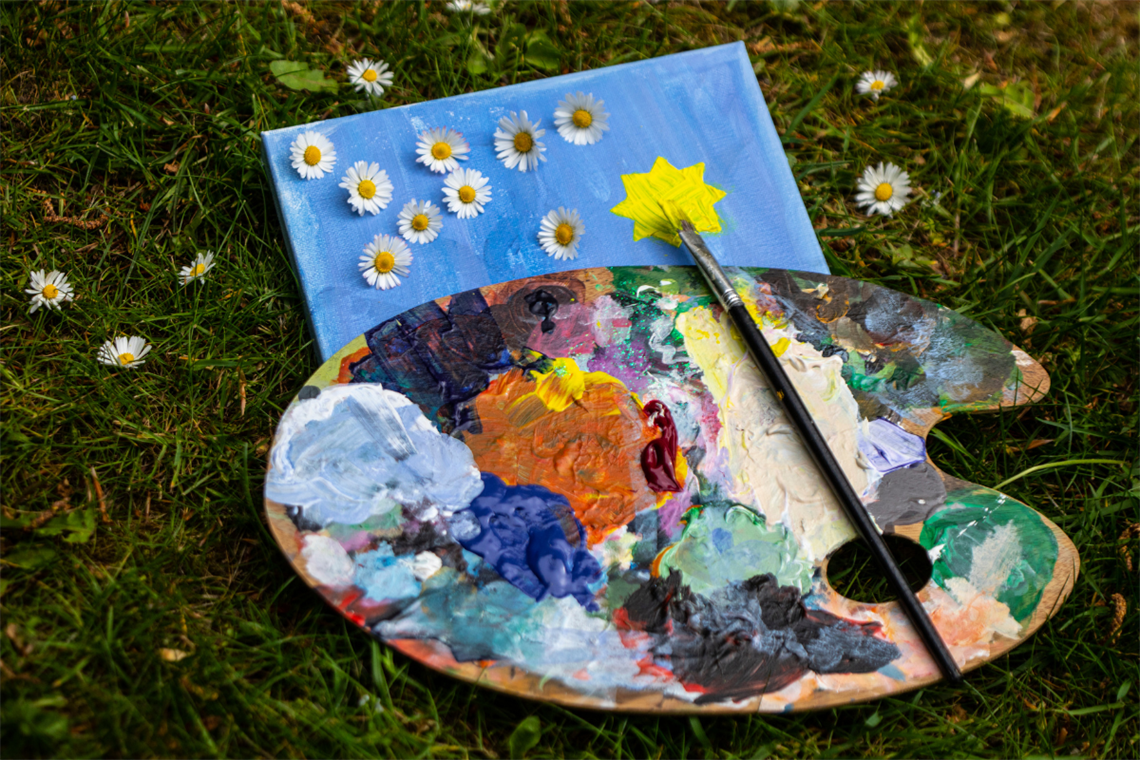 Photo of a wood paint palette with paint, a paint brush laying on grass